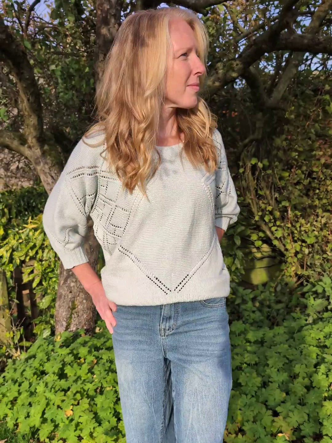 Emma wearing the Tessera sweater in a garden, showing the lace sleeve and eyelet V